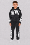 A young boy modeling a black acid-wash hoodie and matching sweatpants set with white flame graphics and 'REBEL' text on the chest, photographed against a neutral gray background.