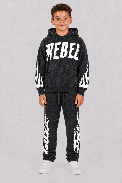 A young boy modeling a black acid-wash hoodie and matching sweatpants set with white flame graphics and 'REBEL' text on the chest, photographed against a neutral gray background.