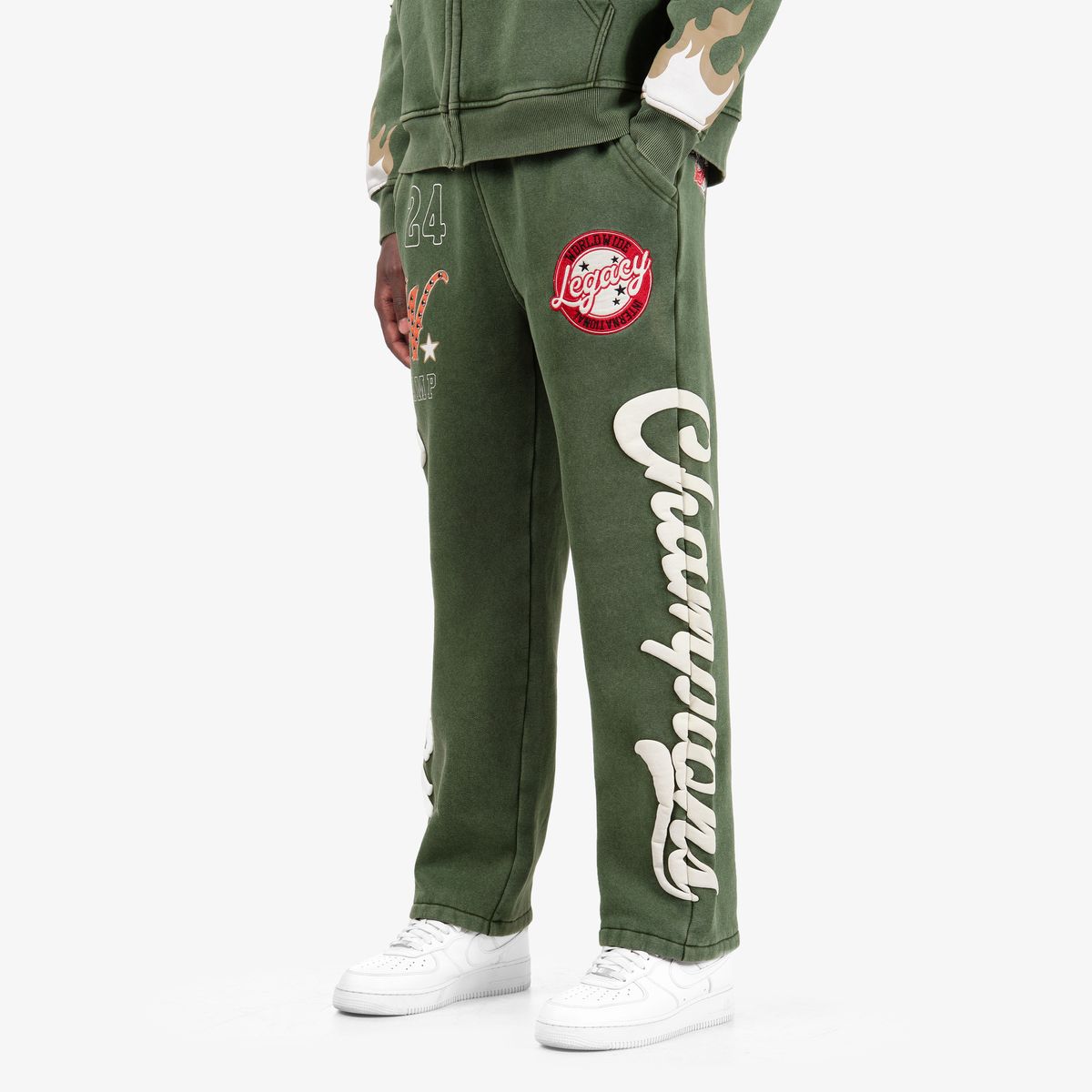 A person wearing olive green Champion sweatpants with a large white script logo down the left leg, a red circular patch on the right thigh, and white sneakers, shown against a plain light gray studio background.