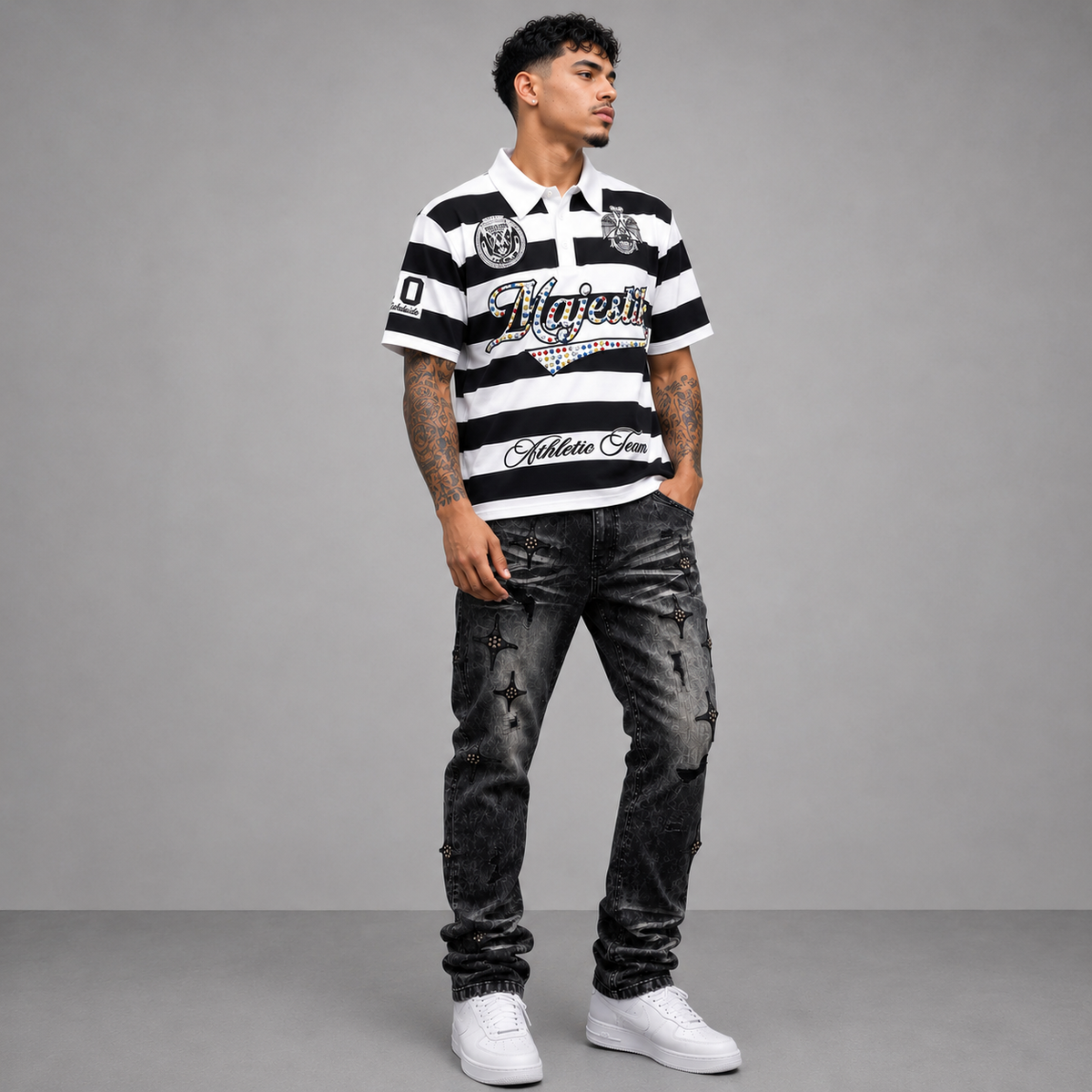 A man modeling a black and white striped polo shirt with 'Vitaforte' and 'Golden Arms' branding, paired with distressed black jeans and white sneakers, against a gray studio background.