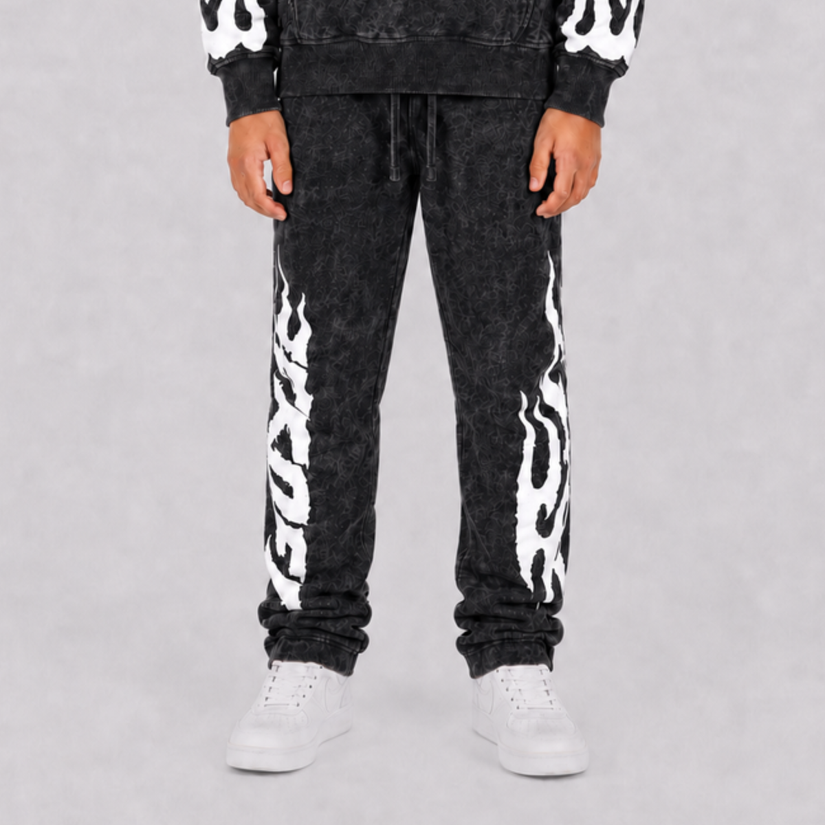 A person wearing black acid-wash sweatpants with bold white flame graphics down the sides, paired with a matching black hoodie and white sneakers, shown from the waist down against a light gray studio background.