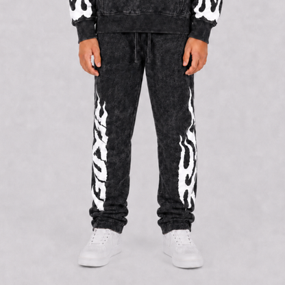 A person wearing black acid-wash sweatpants with bold white flame graphics down the sides, paired with a matching black hoodie and white sneakers, shown from the waist down against a light gray studio background.