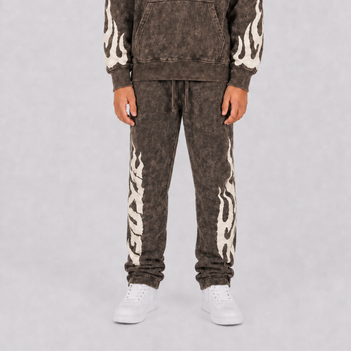 A person wearing a matching acid-wash brown hoodie and jogger sweatpants set with cream flame patterns down the sleeves and legs, paired with white sneakers, against a light gray studio background.