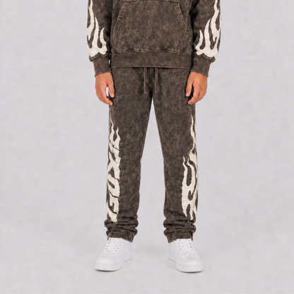 A person wearing a matching acid-wash brown hoodie and jogger sweatpants set with cream flame patterns down the sleeves and legs, paired with white sneakers, against a light gray studio background.