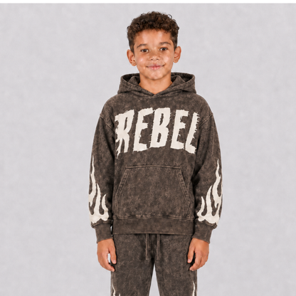 A young boy wearing a dark brown acid-wash hoodie with 'REBEL' printed in distressed white letters across the chest and white flame graphics on the sleeves, paired with matching acid-wash sweatpants, set against a plain gray studio background.